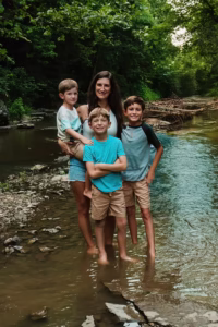 Family standing barefoot in shallow creek surrounded by trees and rocky stream banks