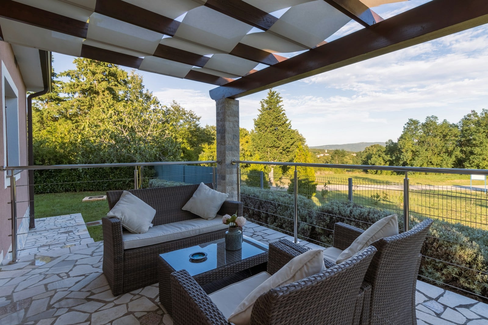 stone patio with wicker seating set under slatted roof and metal railing overlooking green landscape