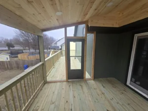 covered elevated deck with wood railing and screen door leading to open outdoor walkway