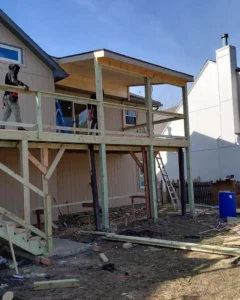 workers building an elevated wooden deck frame with stairs and a partially roofed section attached to a house