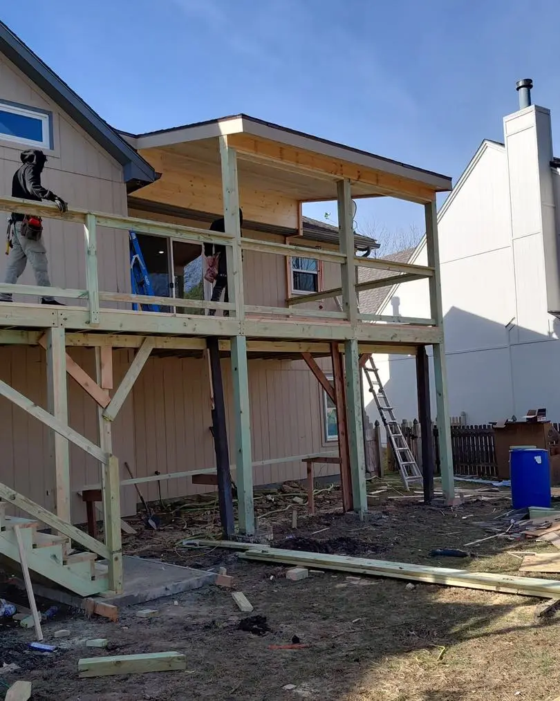workers building an elevated wooden deck frame with stairs and a partially roofed section attached to a house